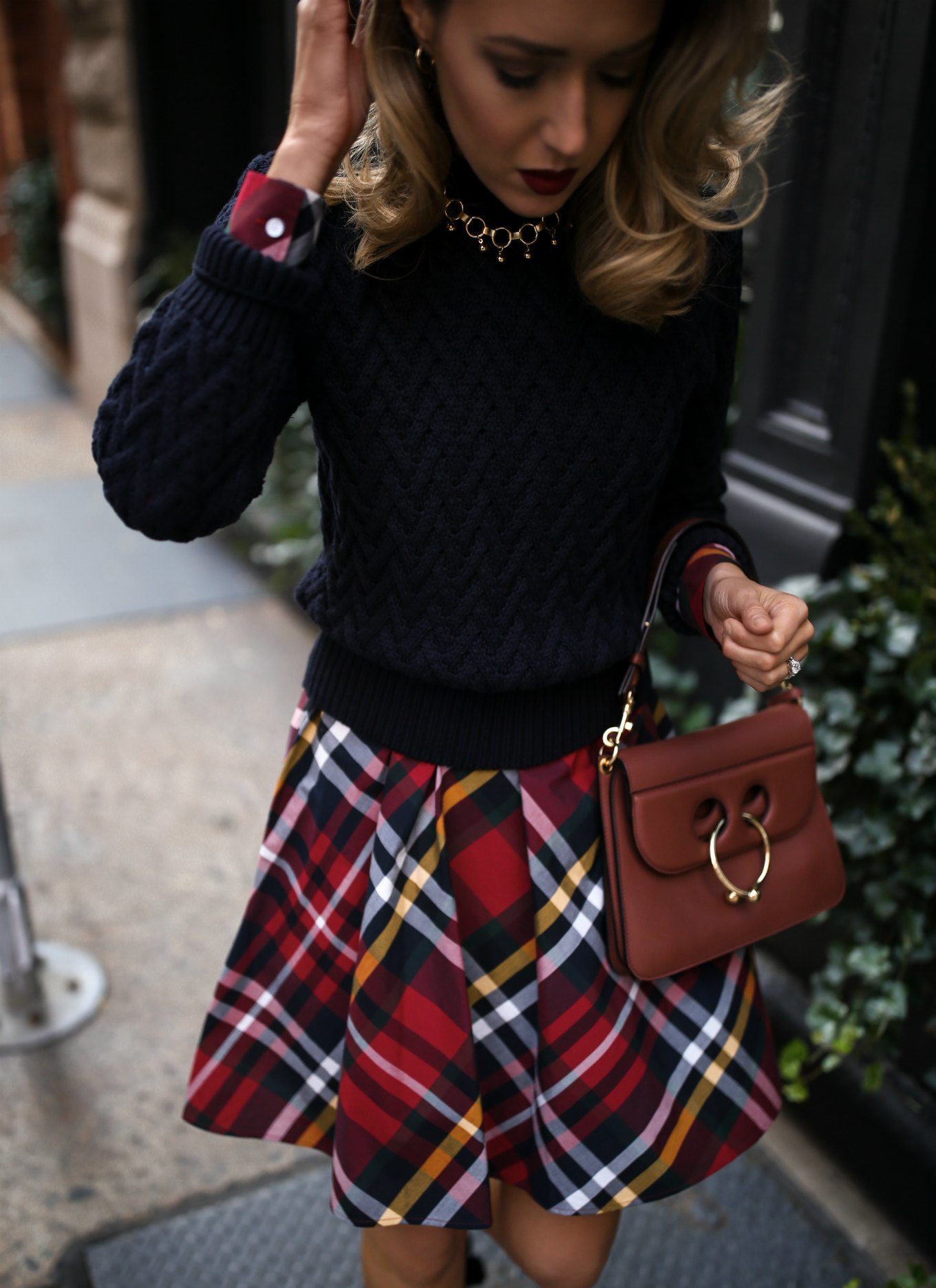 Mary Orton of Memorandum wears a Brooks Brothers red tartan plaid shirt dress with a navy cable knit sweater, a brown leather JW Anderson handbag and a gold statement necklace for a casual office holiday party