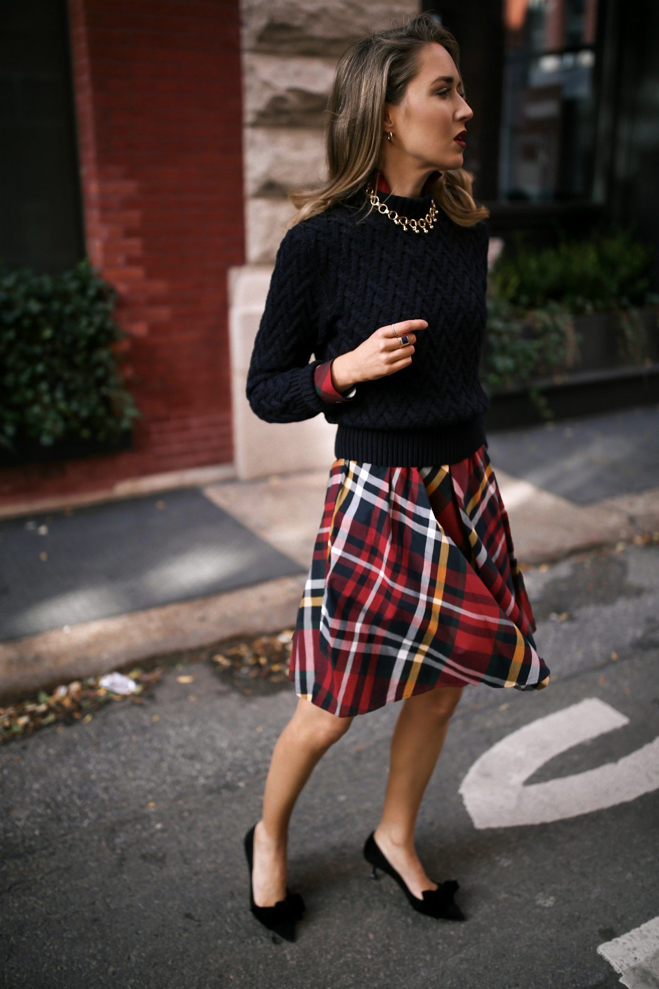 Mary Orton wears a Brooks Brothers red tartan plaid shirt dress with a navy cable knit sweater, a brown leather JW Anderson handbag, black Miu Miu bow pumps and a gold statement necklace for a casual office holiday party