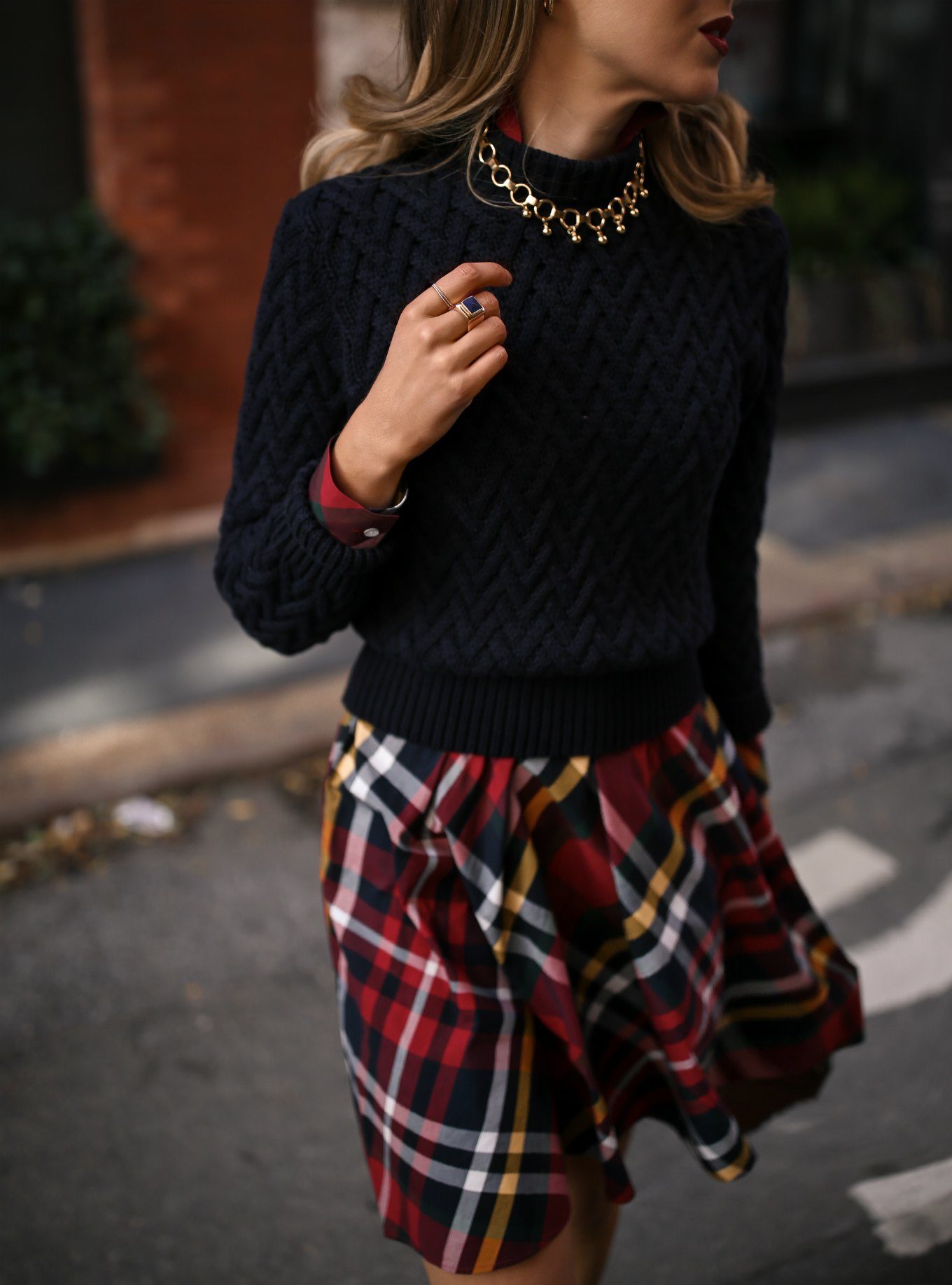 Mary Orton of Memorandum wears a Brooks Brothers red tartan plaid shirt dress with a navy cable knit sweater, a brown leather JW Anderson handbag and a gold statement necklace for a casual office holiday party