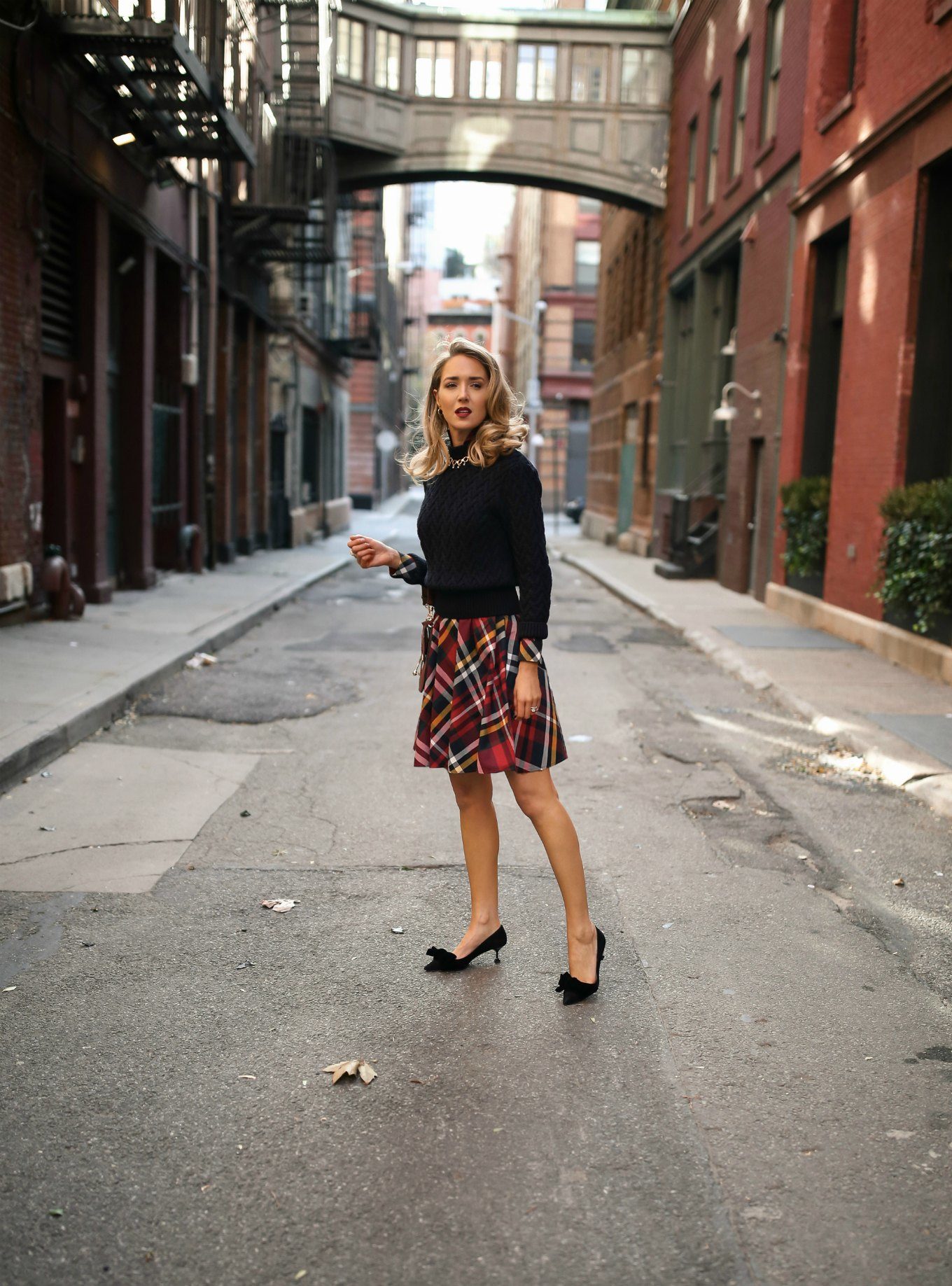 Mary Orton wears a Brooks Brothers red tartan plaid shirt dress with a navy cable knit sweater, a brown leather JW Anderson handbag, black Miu Miu bow pumps and a gold statement necklace for a casual office holiday party