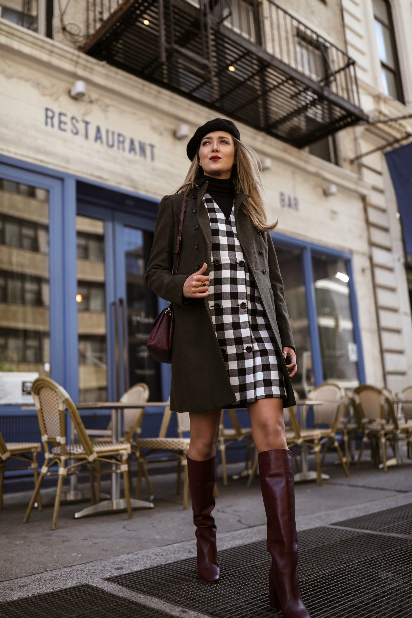 NYC style blogger wearing a gingham pinafore dress over a black turtleneck with red bag, red boots, green coat and black beret